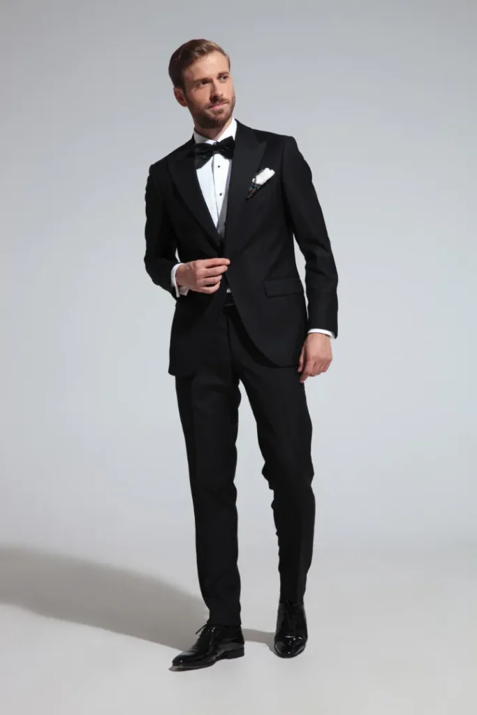 A man on a plain background posing in a black tuxedo